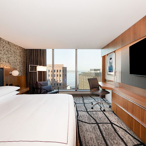 Modern hotel room with a large bed, desk, TV, and a window with a cityscape view. The decor includes wooden accents and patterned wallpaper.