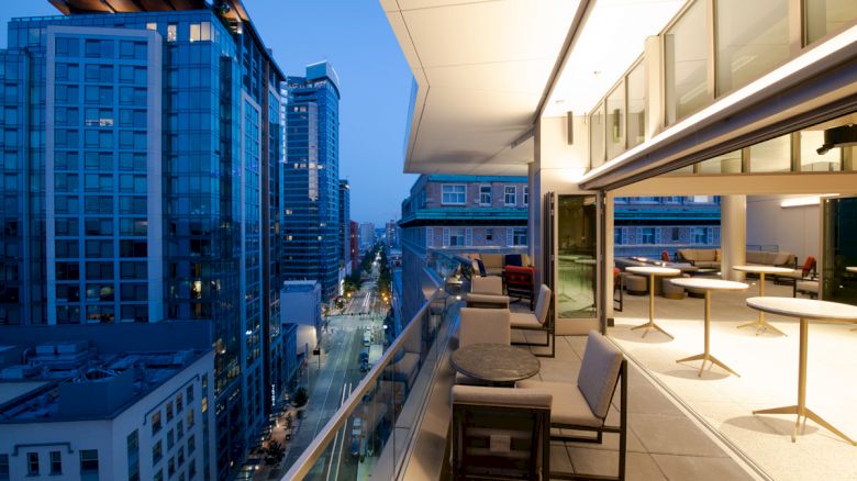 A modern balcony with seating overlooks a city with tall buildings and a street below. The balcony is lit, creating a cozy ambiance.