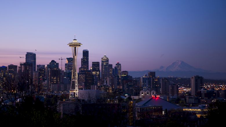 A city skyline at dusk with the Space Needle prominently visible and a mountain range in the background under a clear sky.