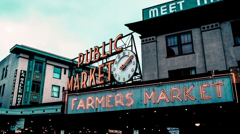 A vibrant sign for a Public Market with a clock and Farmers Market neon lights, flanked by buildings, creating a bustling urban scene.