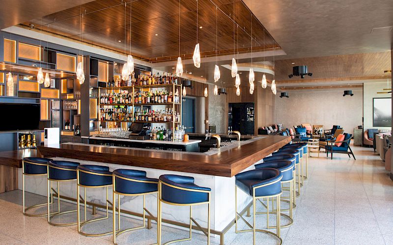 A modern bar with blue stools, hanging lights, and a well-stocked bar counter, featuring a cozy seating area in the background.