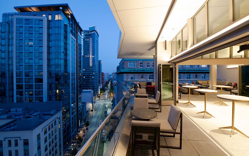 An outdoor terrace at twilight overlooks a street and tall buildings, featuring modern seating and tables, with a glass railing and city view.