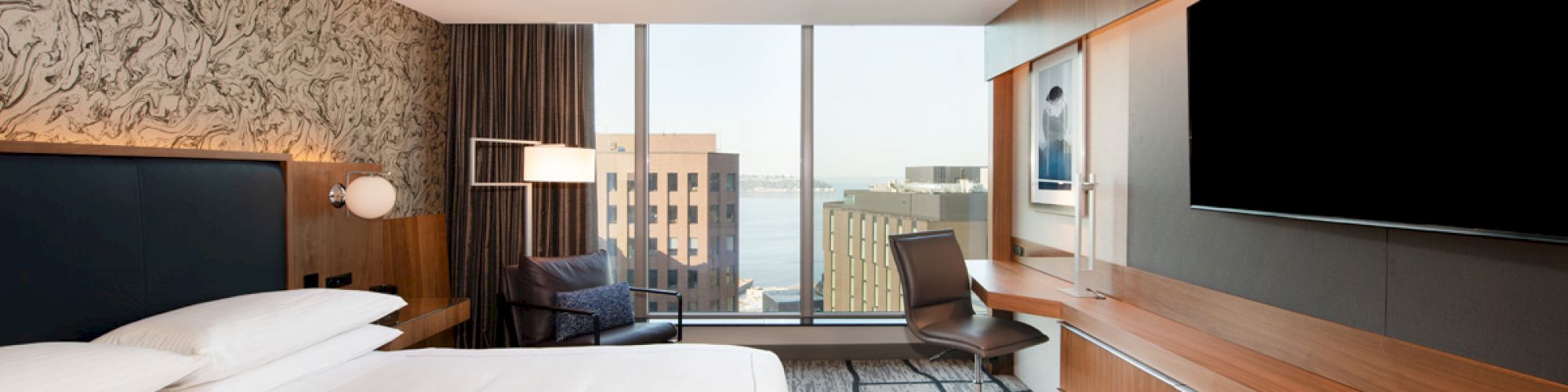 The image shows a modern hotel room with a large bed, a desk with a chair, a flat-screen TV, and a window with a city view.