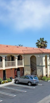 The image shows a two-story motel with a parking lot, several cars parked, palm trees, and a clear blue sky in the background.