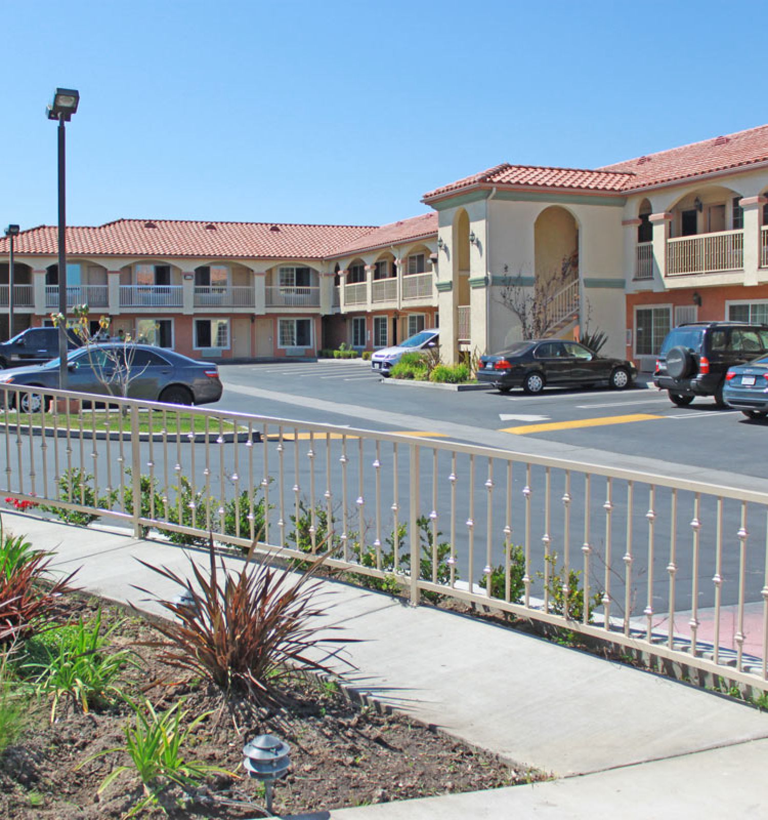 This image shows the exterior of a two-story motel with parked cars, a parking lot, and a sidewalk with plants and a fence in the foreground.