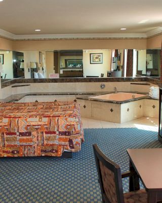 A spacious hotel room with a bed, jacuzzi, mirrored walls, and a table with chairs. The room has a dresser, microwave, and TV.