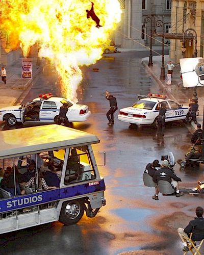 A film set with an explosion, police cars, actors, cameras, and a Universal Studios tour bus in the foreground, with tourists watching the action.