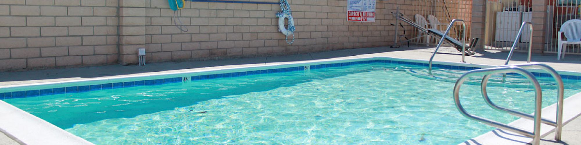 A clean swimming pool with ladders on the side, life ring on the wall, and patio chairs on the poolside, surrounded by a brick wall.